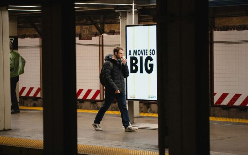 Adult male walking past subway advertisement reading 'A Movie So Big' on urban platform.